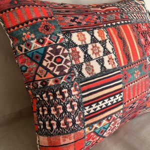 Puede incluir: Un coj&iacute;n cuadrado con un dise&ntilde;o de patchwork. El coj&iacute;n presenta una variedad de patrones geom&eacute;tricos en tonos de rojo, naranja, verde azulado y negro. El coj&iacute;n parece estar hecho de una tela suave.