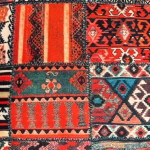 Puede incluir: Primer plano de un textil patchwork con una vibrante mezcla de colores y patrones. El dise&ntilde;o presenta cuadrados con motivos geom&eacute;tricos y tribales en tonos de rojo, naranja, verde azulado, negro y blanco. La tela tiene una apariencia texturizada.