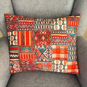 Puede incluir: Coj&iacute;n rectangular decorativo con dise&ntilde;o de patchwork. El coj&iacute;n presenta una variedad de patrones geom&eacute;tricos en tonos rojos, naranjas, negros y blancos. El coj&iacute;n est&aacute; sobre un sill&oacute;n gris.