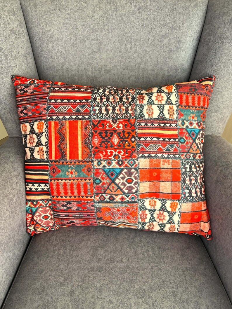 Puede incluir: Coj&iacute;n rectangular decorativo con dise&ntilde;o de patchwork. El coj&iacute;n presenta una variedad de patrones geom&eacute;tricos en tonos rojos, naranjas, negros y blancos. El coj&iacute;n est&aacute; sobre un sill&oacute;n gris.
