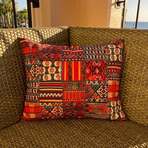 Puede incluir: Un coj&iacute;n cuadrado decorativo con dise&ntilde;o de patchwork. Presenta diversos patrones en tonos rojos, naranjas, negros y turquesas. Se encuentra sobre una silla de mimbre trenzado, sugiriendo uso en exteriores.