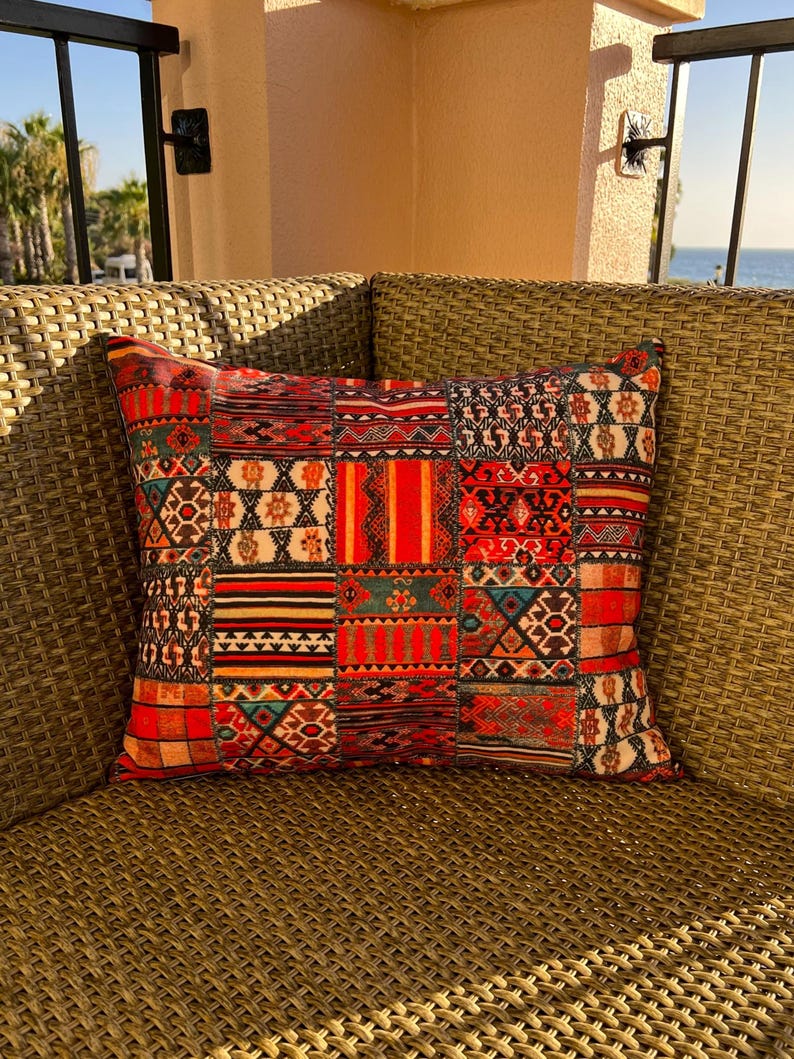 Puede incluir: Un coj&iacute;n cuadrado decorativo con dise&ntilde;o de patchwork. Presenta diversos patrones en tonos rojos, naranjas, negros y turquesas. Se encuentra sobre una silla de mimbre trenzado, sugiriendo uso en exteriores.