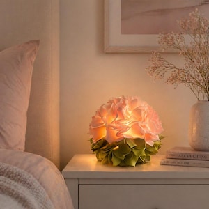 May include: A spherical table lamp featuring layered, petal-like fabric elements in soft pink at the top and muted green at the base. The lamp emits a warm, diffused glow from within, highlighting the texture of the overlapping fabric layers.