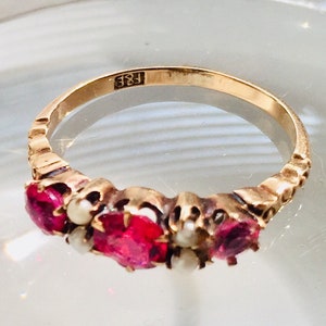 Peut inclure: Une bague en or avec une bande décorée d'un motif répétitif. La bague est sertie de trois pierres précieuses rouges et de deux perles blanches. La bague est marquée "18E".