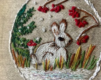 May include: Embroidered artwork featuring a white and brown rabbit in a natural setting. The scene includes green foliage, a red mushroom, and red berries. The artwork is framed with a white border and stitched on a beige fabric.