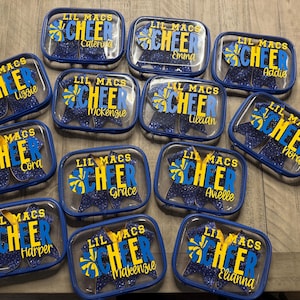 May include: Clear rectangular cases with blue trim, each labeled "Lil Macs Cheer" with a name. The cases contain blue star-shaped items. The text is in yellow and blue.