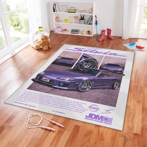 May include: A rectangular rug featuring a purple Nissan Silvia S15 car with chrome wheels. The rug has text that reads "Nissan Silvia" and "JDM Japanese Domestic Market". A jump rope lies on the wooden floor in front of the rug.