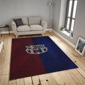 Könnte beinhalten: Ein rechteckiger Teppich mit einem rot-blauen Farbblock-Design. Der Teppich hat ein silberfarbenes Emblem in der Mitte. Der Teppich liegt auf einem Holzboden, mit einem grauen Sofa und einem Fenster im Hintergrund.