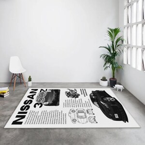 以下が含まれることがあります： 「NISSAN 350Z」の文字が入った、白地に黒と白のデザインの長方形のラグ。車のイラストとスペイン語のテキストが含まれています。ラグは灰色の床に置かれています。