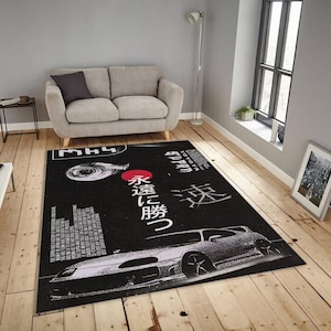 May include: Black rectangular area rug featuring a white car graphic and Japanese text. The rug has a red sun graphic and a white turbocharger graphic. The rug is placed on a wooden floor in a room with a gray sofa.