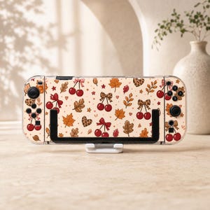 May include: A handheld gaming console featuring a cream-coloured skin with a repeating pattern of red cherries, brown autumn leaves, leopard print hearts, and small bows. The device is held in a black plastic stand and has clear protective casing.