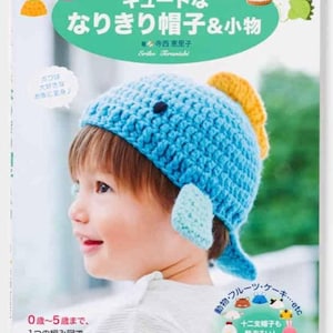 以下が含まれることがあります： 青い魚の帽子をかぶった子供が表紙の書籍。タイトルは「かぎ針編み キュートな なりきり帽子＆小物」と日本語で書かれています。様々な帽子やアクセサリーのイラストも含まれています。