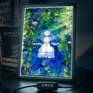 May include: A framed anime art piece featuring a serene scene with a character in a white dress amidst lush greenery and a blue water setting. The frame has a black border and is illuminated with a soft glow. A control panel with buttons is visible.