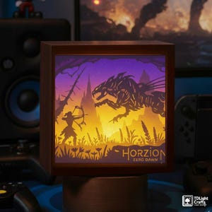 Könnte beinhalten: Ein Holz-Leuchtkasten mit einem geschichteten Papier-Schnitt-Design aus dem Videospiel "Horizon Zero Dawn". Das Kunstwerk zeigt einen silhouettierten Bogenschützen, ein mechanisches Tier und den Titel des Spiels vor einem lila-gelben Farbverlauf.
