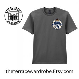 May include: Dark gray t-shirt with a circular emblem featuring a crown and the words "LOS BLANCOS" in navy blue and gold. The shirt also has the text "19 02". The Terrace Wardrobe logo is on the left side of the shirt.