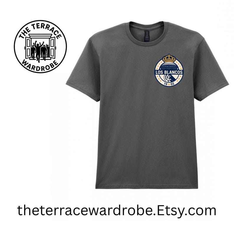 May include: Dark gray t-shirt with a circular emblem featuring a crown and the words "LOS BLANCOS" in navy blue and gold. The shirt also has the text "19 02". The Terrace Wardrobe logo is on the left side of the shirt.