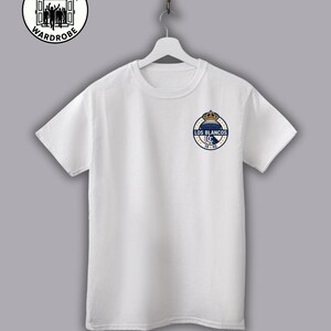 May include: White crew neck t-shirt with a small, circular emblem on the left chest. The emblem features a crown above the words "LOS BLANCOS" in a navy blue and gold design. The Terrace Wardrobe logo is at the top left.