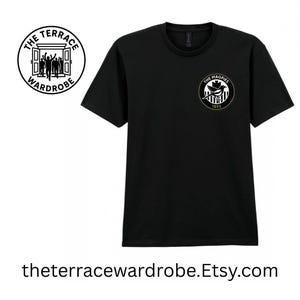 May include: Black t-shirt with two circular logos. One logo reads "The Terrace Wardrobe" and features a doorway with silhouettes. The other logo says "The Magpies 1892" with a bird and striped design. The website address is also visible.