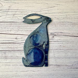 May include: A flat ceramic wall hanging shaped like a sitting hare. The piece features a deep blue glaze with darker circular patterns and a textured, speckled grey border. The hare has one long, pointed ear with a cream-coloured centre detail.