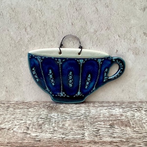 Könnte beinhalten: Wandbehang in Form einer Keramik-Teetasse. Die Tasse ist tiefblau mit weißem Rand und Henkel. Das Design zeigt ein Blumenmuster in einem helleren Blau. Ein schwarzer Drahtgriff ist zum Aufhängen angebracht.