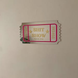 May include: A mirrored ticket-shaped wall decor with the words "SHIT SHOW" in gold lettering, framed by a pink border. The ticket also includes the words "Admit One" and the numbers 193405 and 135410.