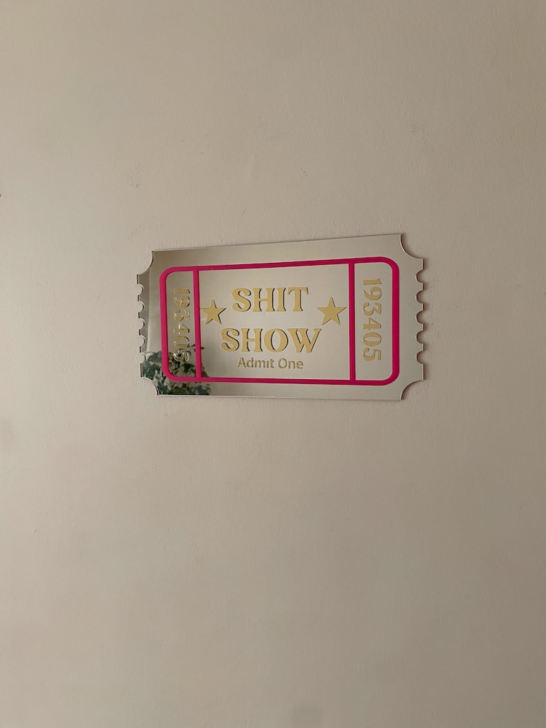 May include: A mirrored ticket-shaped wall decor with the words "SHIT SHOW" in gold lettering, framed by a pink border. The ticket also includes the words "Admit One" and the numbers 193405 and 135410.