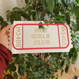 May include: A white sign with a red border and the words "HOT GIRLS CLUB" in gold lettering. The sign has the numbers "193405" on each end. The sign is held in front of a green leafy plant.