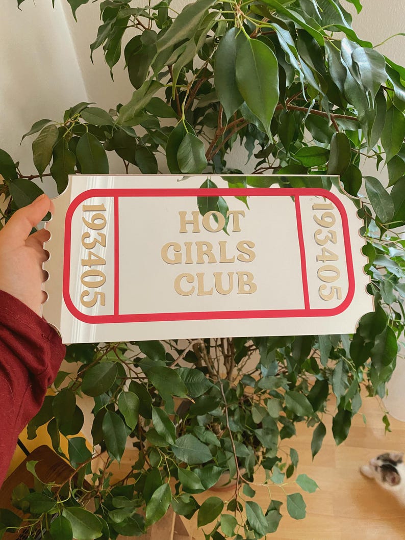 May include: A white sign with a red border and the words "HOT GIRLS CLUB" in gold lettering. The sign has the numbers "193405" on each end. The sign is held in front of a green leafy plant.