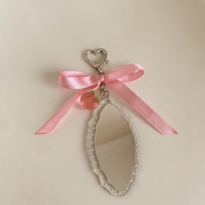 Puede incluir: Un pequeño llavero con espejo ovalado transparente con borde decorativo. El llavero presenta una cinta rosa atada en un lazo, un cierre en forma de corazón y un dije de fresa rosa. El espejo mide aproximadamente 5 cm de largo.