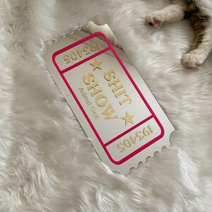 May include: A white ticket with a pink border and gold text that reads "SHIT SHOW" and "Admit One". The ticket has a serrated edge and the number "193405" is printed on it. The ticket is on a white, fluffy surface.
