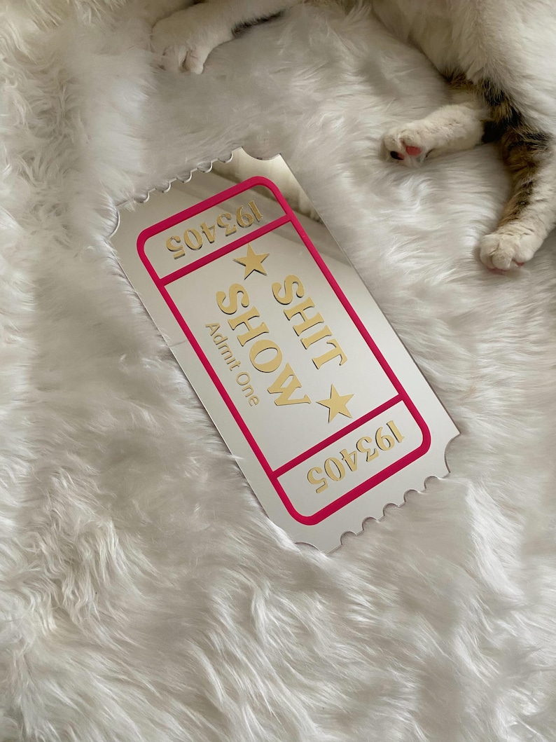 May include: A white ticket with a pink border and gold text that reads "SHIT SHOW" and "Admit One". The ticket has a serrated edge and the number "193405" is printed on it. The ticket is on a white, fluffy surface.