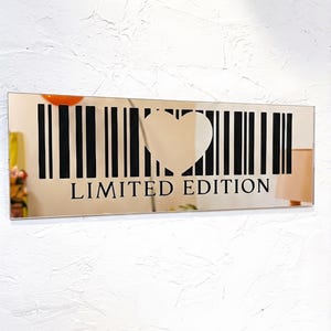 May include: A rectangular mirror with a heart-shaped cutout and a barcode design. The words "LIMITED EDITION" are printed in black below the barcode. The mirror is mounted on a textured white wall.