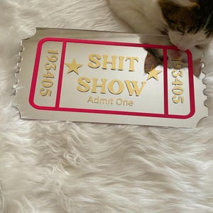 May include: A rectangular mirror with a pink border and the words "SHIT SHOW" and "Admit One" in gold lettering. The mirror has a ticket-like design with the numbers "193405" on each side. A cat is partially visible in the image.