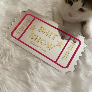 May include: A rectangular, silver-colored ticket with the words "SHIT SHOW" in gold lettering, framed by a pink border. The ticket also features the text "Admit One" and the numbers "193405". A kitten is in the background.