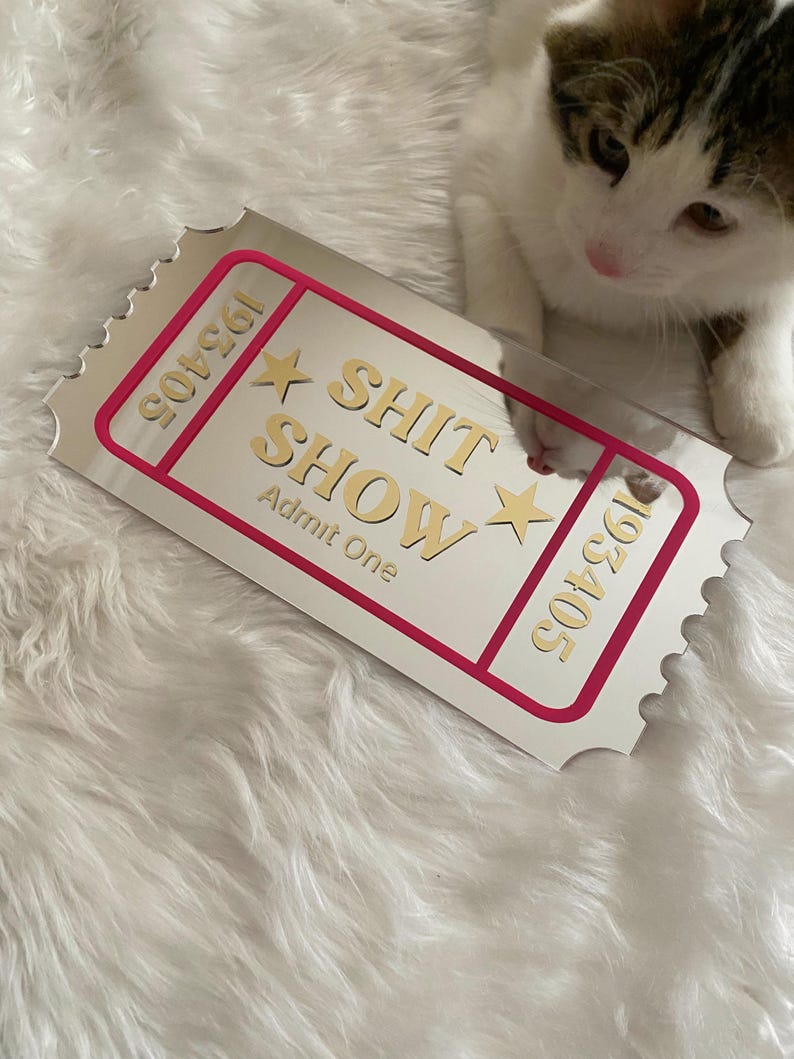May include: A rectangular, silver-colored ticket with the words "SHIT SHOW" in gold lettering, framed by a pink border. The ticket also features the text "Admit One" and the numbers "193405". A kitten is in the background.