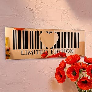 May include: A rectangular gold mirror with a barcode design and a heart-shaped cutout. The words "LIMITED EDITION" are printed below the barcode. Red flowers in a white vase are in the foreground.