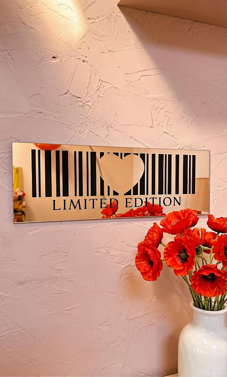 May include: A rectangular gold mirror with a barcode design and a heart-shaped cutout. The words "LIMITED EDITION" are printed below the barcode. Red flowers in a white vase are in the foreground.