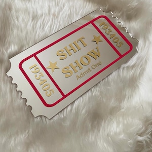 May include: A silver-colored ticket-shaped mirror with a red border and the words "SHIT SHOW" in gold lettering. The ticket also has the numbers "193405" and "Admit One" printed on it. The mirror is on a white, fluffy surface.