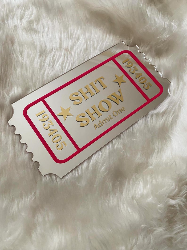 May include: A silver-colored ticket-shaped mirror with a red border and the words "SHIT SHOW" in gold lettering. The ticket also has the numbers "193405" and "Admit One" printed on it. The mirror is on a white, fluffy surface.