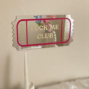 May include: A mirrored ticket-shaped wall decor with the text "FUCK ME CLUB" in gold lettering. The ticket has a pink border and the numbers "193405" on each side. A white candle sits in a decorative holder below.