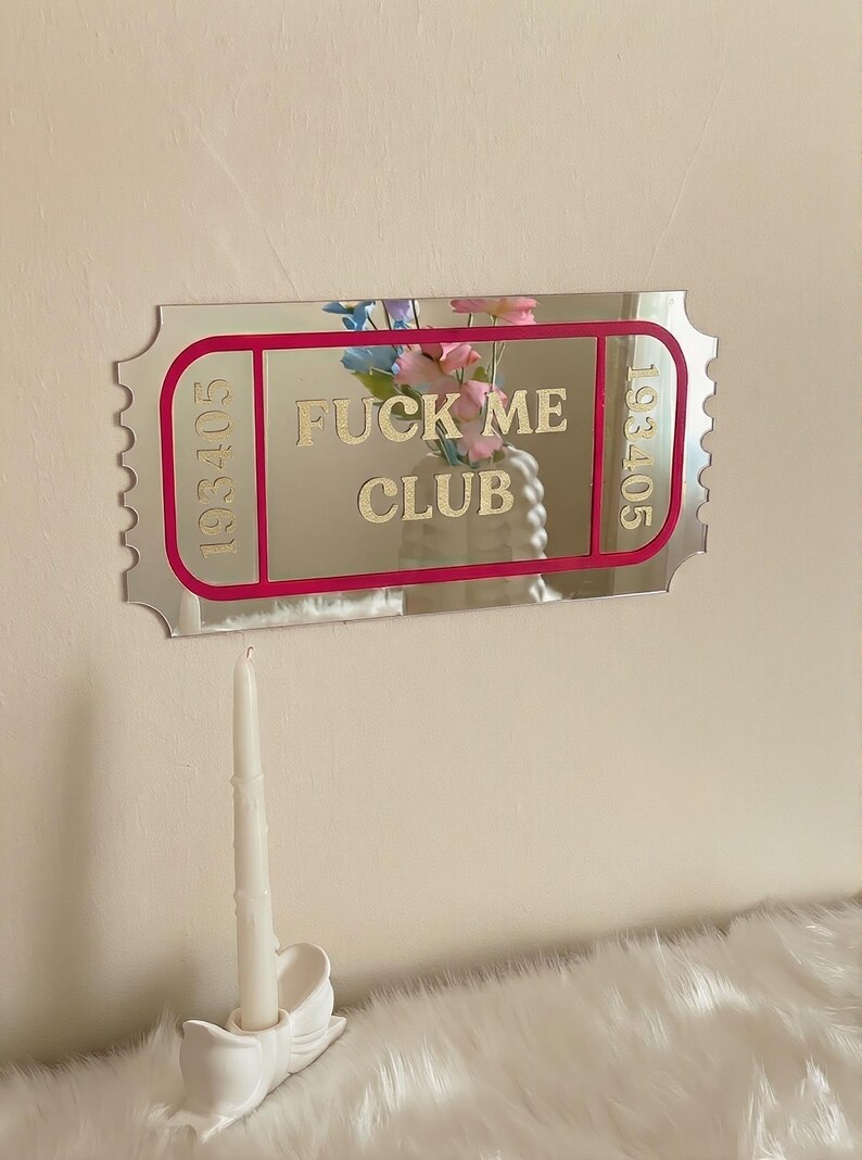 May include: A mirrored ticket-shaped wall decor with the text "FUCK ME CLUB" in gold lettering. The ticket has a pink border and the numbers "193405" on each side. A white candle sits in a decorative holder below.
