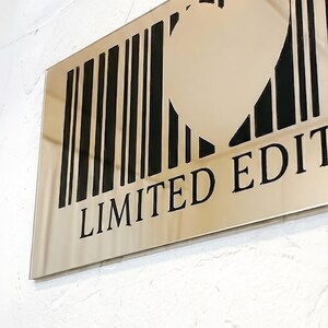 May include: A rectangular, metallic sign with a heart-shaped cutout within a barcode design. The words "LIMITED EDITION" are printed in black below the barcode. The sign is mounted on a textured white wall.