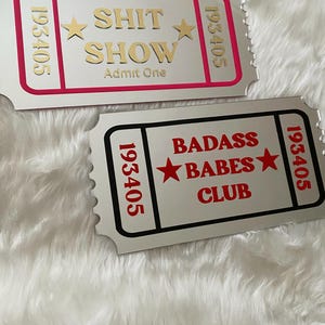 May include: Two silver-colored ticket-shaped signs. One has a pink border and gold text that reads "SHIT SHOW Admit One" with star accents. The other has a black border and red text that reads "BADASS BABES CLUB" with star accents.