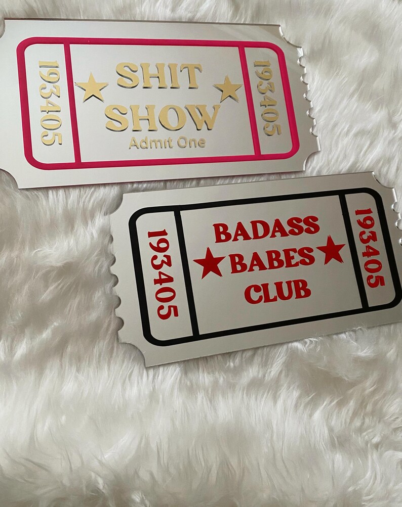 May include: Two silver-colored ticket-shaped signs. One has a pink border and gold text that reads "SHIT SHOW Admit One" with star accents. The other has a black border and red text that reads "BADASS BABES CLUB" with star accents.