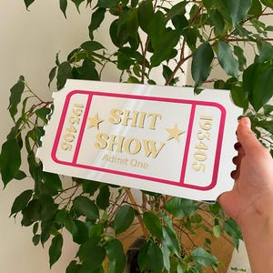 May include: A white ticket-shaped sign with a pink border and gold lettering that reads "SHIT SHOW" and "Admit One". The sign also has gold stars and the numbers "193405" on each side. The sign is held in front of a green leafy plant.