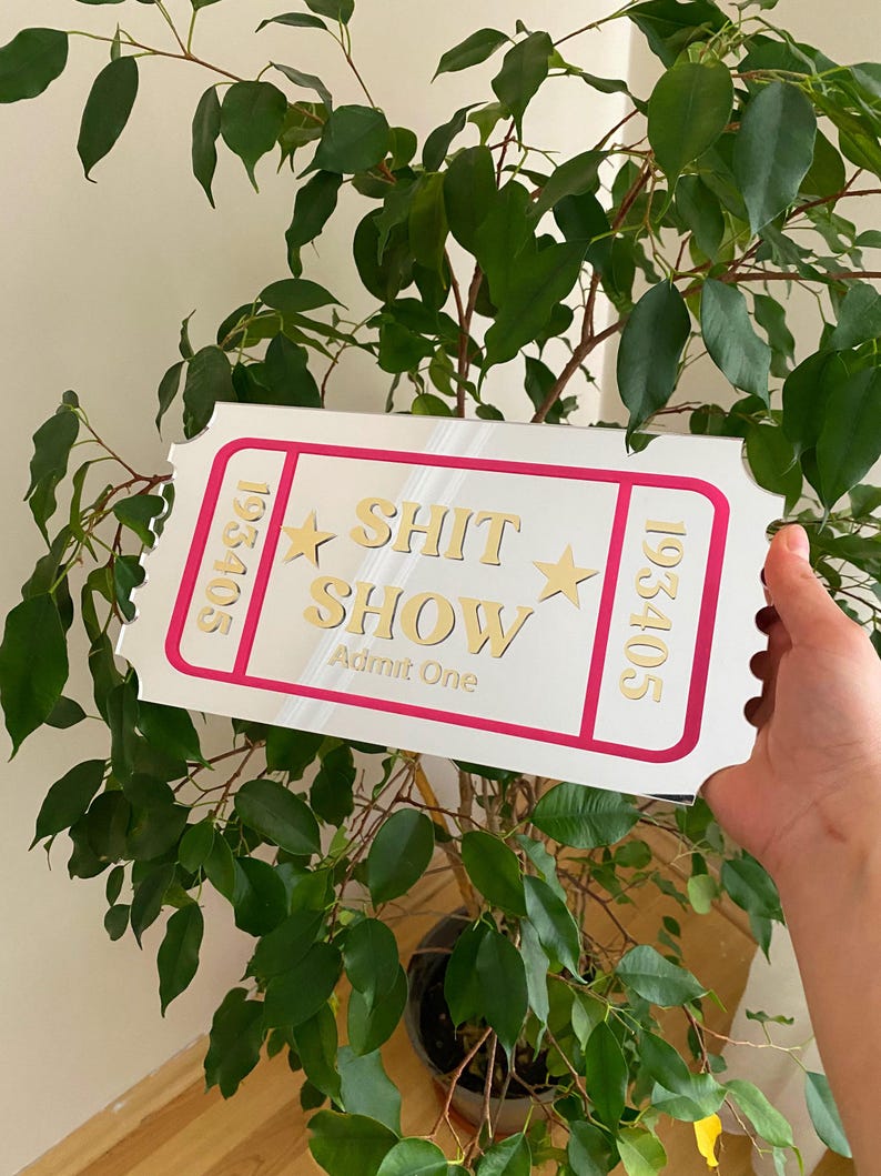 May include: A white ticket-shaped sign with a pink border and gold lettering that reads "SHIT SHOW" and "Admit One". The sign also has gold stars and the numbers "193405" on each side. The sign is held in front of a green leafy plant.