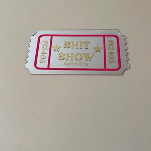 May include: A silver-colored ticket with the words "SHIT SHOW" in gold lettering, framed by a pink border. The ticket also includes the text "Admit One" and the numbers "193405".
