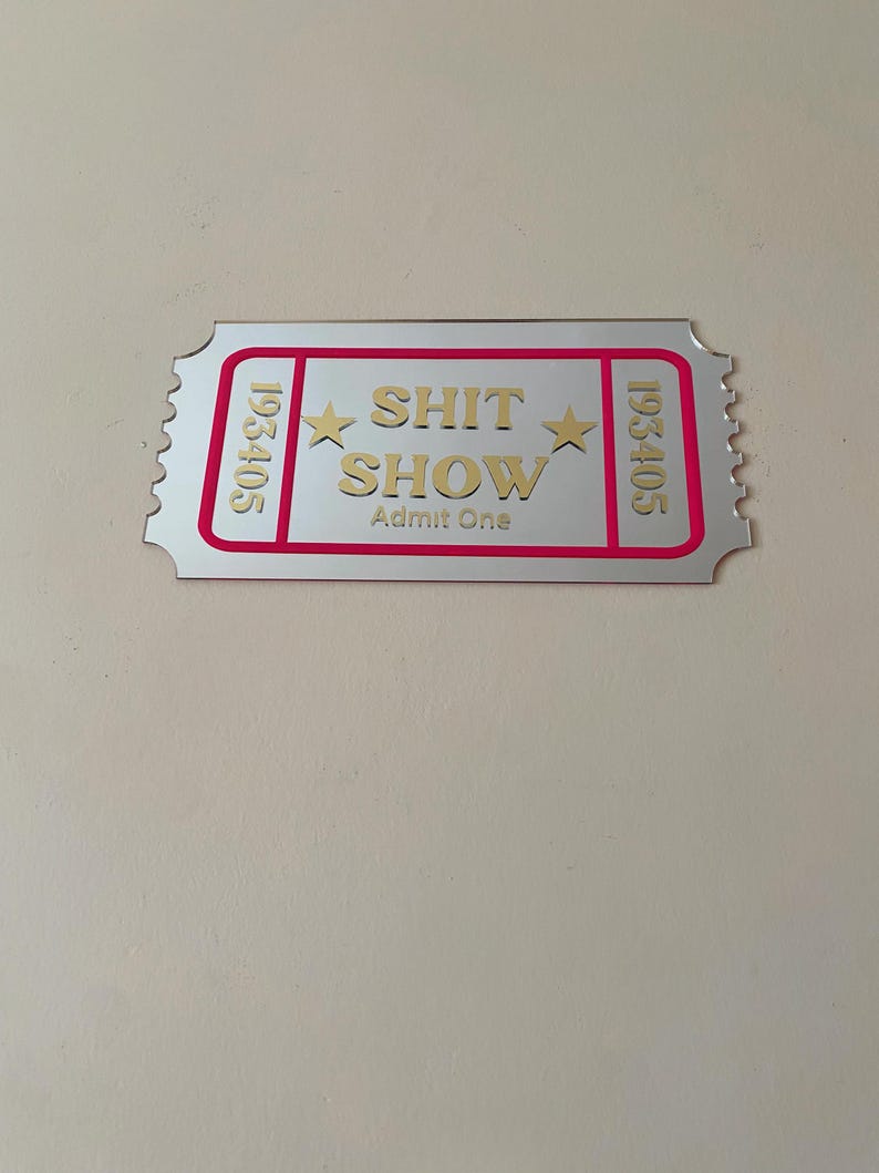 May include: A silver-colored ticket with the words "SHIT SHOW" in gold lettering, framed by a pink border. The ticket also includes the text "Admit One" and the numbers "193405".