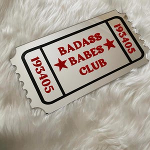 May include: A rectangular ticket-shaped mirror with the words "BADASS BABES CLUB" in red, along with two red stars. The ticket has the number "193405" in red on each end, and is framed with a black border.