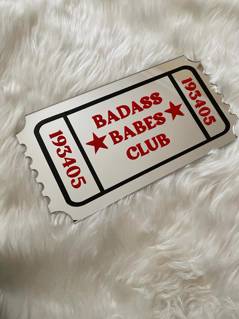 May include: A rectangular ticket-shaped mirror with the words "BADASS BABES CLUB" in red, along with two red stars. The ticket has the number "193405" in red on each end, and is framed with a black border.